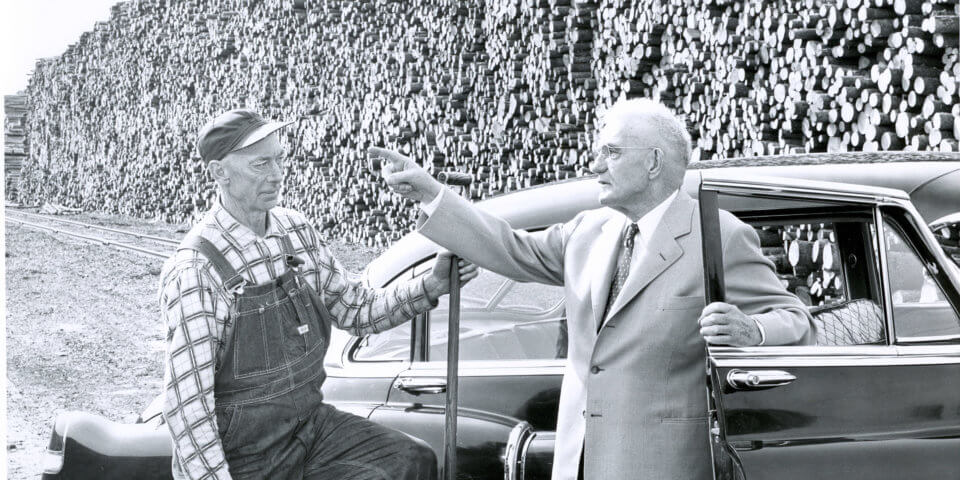 CK by car talking to man in overalls log pile in bkgd 1950.364.10001